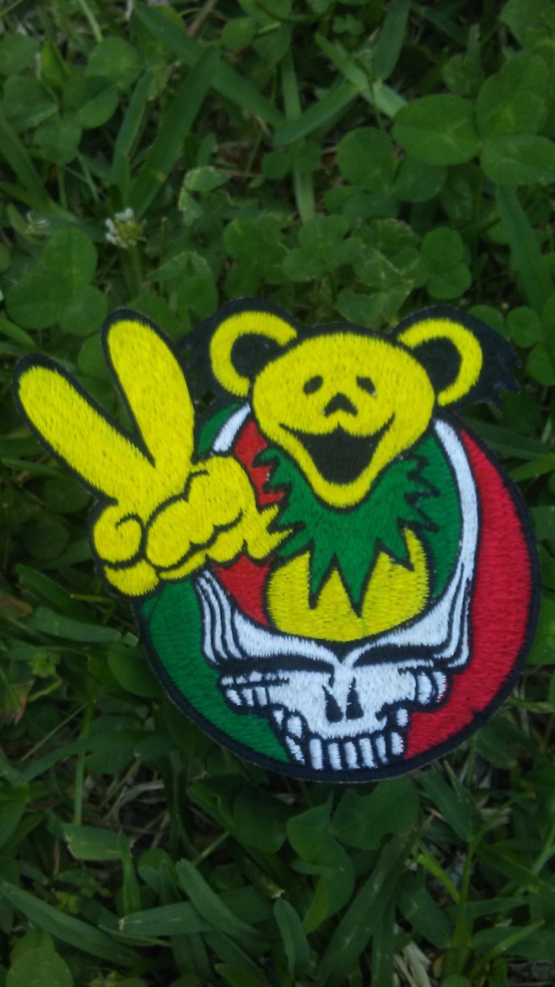 May include: Embroidered patch with a yellow bear peace sign, and a red, green, and white skull design. The patch is set against a background of green grass and clover.