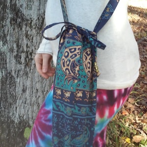 May include: A blue and green patterned drawstring bag with a long strap worn over the shoulder. The bag is made of fabric and has a tassel.