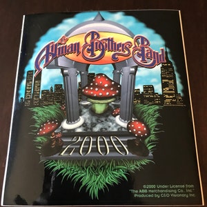 May include: A black and green sticker with a mushroom and a cityscape in the background. The text "The Allman Brothers Band" and "2000" are visible. The sticker has a glossy finish.
