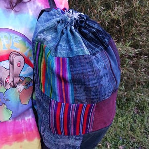 May include: A patchwork backpack with a drawstring closure. The bag features a variety of blue denim and patterned fabric patches, with accents of red, purple, and green stripes. The bottom is a dark green color.