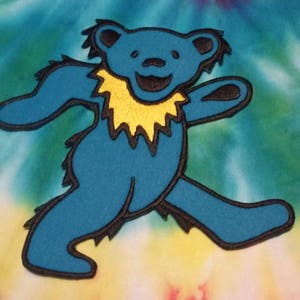 May include: A blue tie-dye fabric with a large, blue, smiling bear patch with a yellow collar. The bear is walking with its arms outstretched.