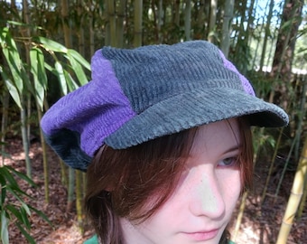 May include: A purple and dark gray corduroy newsboy cap with a wide brim. The cap is worn by a person with long brown hair.