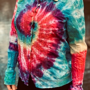 May include: A long-sleeved button-down shirt with a tie-dye pattern in shades of blue, red, purple, and yellow. The shirt has a white collar and a lace trim around the neckline and cuffs.