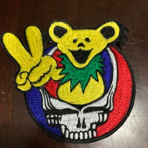 May include: A yellow and green smiling bear with a peace sign and a skull and crossbones design. The bear is wearing a green lei. The skull and crossbones are in a circle with red, blue, and white colours.