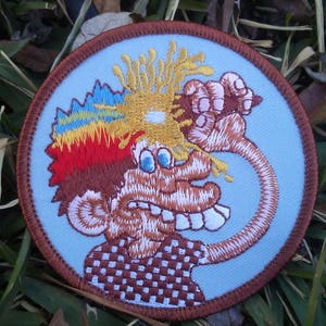 May include: Embroidered patch with a cartoon character design. The patch has a light blue background and a brown border. The character has wild hair in yellow, red, and blue, and a checkered shirt. The patch is circular.