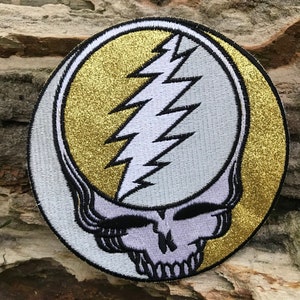 May include: Circular embroidered patch with a skull and lightning bolt design. The patch has a gold glitter background and a white skull with black detailing. The patch is approximately 7.6 cm in diameter.
