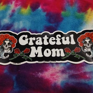 May include: A sticker with the words "Grateful Mom" in white, flanked by skull illustrations with red flower crowns and roses. The sticker has a black border and is set against a tie-dye background.