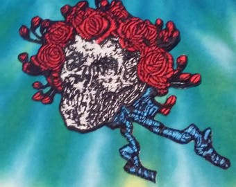 Könnte beinhalten: Gestickter Aufnäher mit einem Totenkopf, einer Krone aus roten Rosen und einem blauen Band. Der Aufnäher befindet sich auf einem tie-dye-Hintergrund.