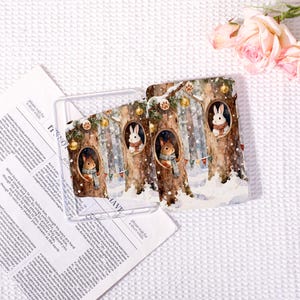 May include: Clear phone case featuring a winter forest scene with rabbits in tree hollows. The design includes snow, garlands, and ornaments. The case is made of clear plastic and is designed to protect a phone.