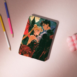 Può includere: Una custodia per telefono rettangolare con un'illustrazione anime gotica. L'opera raffigura una donna bionda in abito nero e un uomo con corna rosse, sullo sfondo di una luna piena e un castello. La custodia ha una finitura lucida.