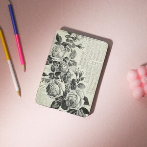 Peut inclure: Une coque de téléphone rectangulaire avec un motif floral de roses en noir et blanc. Le motif comprend un fond d'impression de journal vintage. L'étui est posé sur une surface rose avec deux crayons et une bougie rose.