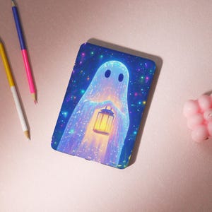 Może przedstawiać: Niebieskie etui na tablet z świecącym duchem trzymającym latarnię. Duch otoczony jest gwiaździstym niebem z kolorowymi plamkami. Etui leży na różowej powierzchni z ołówkami i różową świecą.
