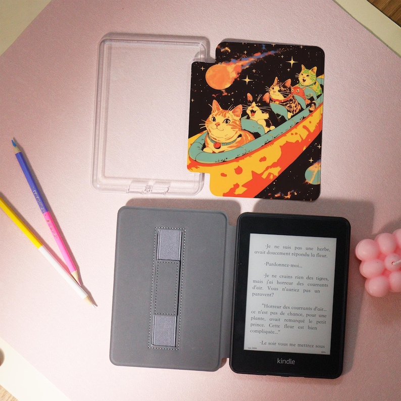Peut inclure: Un lecteur de livres &eacute;lectroniques Kindle noir avec une housse grise et un &eacute;tui en plastique transparent. Une couverture d&eacute;corative avec un motif de chats spatiaux est &agrave; c&ocirc;t&eacute; de l'&eacute;tui. Le Kindle affiche du texte en fran&ccedil;ais.