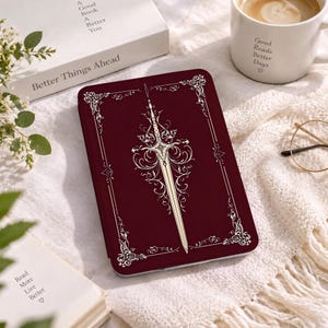 May include: A rectangular dark red e-reader case featuring a central cream-colored ornate sword illustration with intricate filigree detailing and a decorative border frame, presented in a flat orientation on a textured white fabric surface.