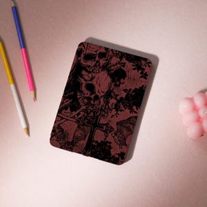 Può includere: Una custodia rettangolare bordeaux con un design a teschio e ossa incrociate nere. Il design include motivi floreali e di pipistrelli. La custodia è su una superficie rosa con matite e una candela rosa.
