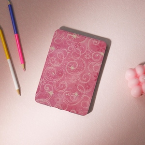May include: A pink rectangular case with a swirling, glittery design. Three coloured pencils and a small pink candle are also visible. The pencils are yellow, blue, and pink.