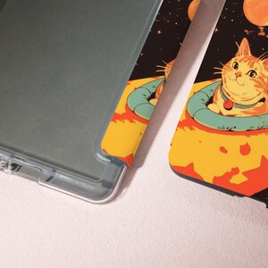 Peut inclure: Un &eacute;tui de t&eacute;l&eacute;phone transparent &agrave; c&ocirc;t&eacute; d'un &eacute;tui de t&eacute;l&eacute;phone avec une illustration color&eacute;e d'un chat en combinaison spatiale. Le chat est orange et blanc, avec un collier bleu et une &eacute;tiquette rouge. Le fond est noir avec des plan&egrave;tes et des &eacute;toiles.