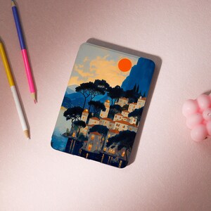 May include: A rectangular case featuring a coastal town scene with a vibrant orange sun, blue mountains, and buildings with warm-toned roofs. Dark green trees and the sea complete the landscape. The case is likely for a tablet or phone.