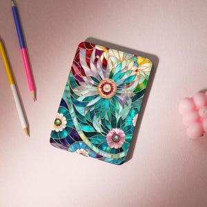 Può includere: Un design floreale rettangolare e colorato. Il design presenta un grande fiore centrale con petali turchese, blu e verdi, circondato da fiori più piccoli. Il design ha un effetto vetrata con contorni dorati. Matite e una candela rosa sono sullo sfondo.