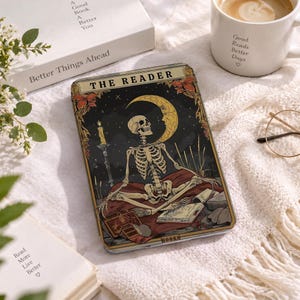 May include: A rectangular e-reader case featuring a graphic illustration of a skeleton sitting in a meditative pose under a crescent moon. The design includes a lit candle, open books, and the text THE READER and BOOKS in a gold-toned frame.