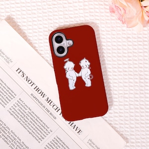 May include: A burgundy phone case featuring a white illustration of two cherubic figures holding hands. One figure has a halo, the other a devil's tail. The case has a camera cutout and the text "IT'S NOT HOW MUCH YOU HAVE" is visible.