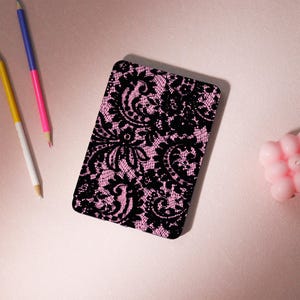 Op de afbeelding: Een rechthoekig item met een zwart kanten patroon op een roze achtergrond, mogelijk een telefoonhoesje of notitieboek. Het ligt op een roze oppervlak, met kleurpotloden en een kleine roze kaars in de buurt.