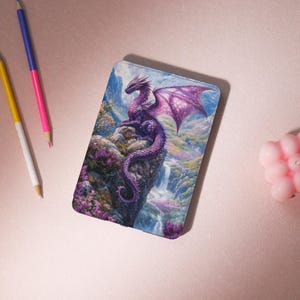 May include: A tablet case with a purple dragon design, sitting on a cliff with a waterfall backdrop. The dragon has detailed wings and is set against a mountain and foliage scene. The case is rectangular with rounded corners.