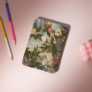 May include: A rectangular case featuring a floral painting with white, pink, and beige flowers, green leaves, and a blue sky background. The case is on a pink surface with three pencils and a pink candle.
