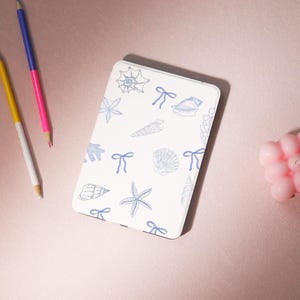 May include: White e-reader cover with a blue and white seashell and starfish pattern. The cover has a blue ribbon design.