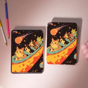 Peut inclure: Deux &eacute;tuis rectangulaires noirs avec une illustration color&eacute;e de chats dans un vaisseau spatial. Les chats sont dans un vaisseau spatial bleu et jaune avec une com&egrave;te rouge et orange en arri&egrave;re-plan. Le fond est noir avec des &eacute;toiles.