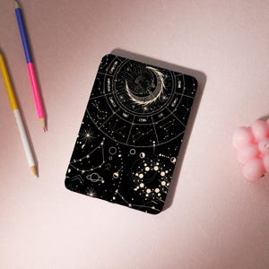 Könnte beinhalten: Schwarzes rechteckiges Notizbuch mit einem Himmelsdesign mit Tierkreiszeichen, Sternbildern und einem Halbmond. Das Notizbuch hat abgerundete Ecken. Stifte in Gelb, Rosa und Blau sind im Hintergrund.