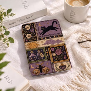 May include: A rectangular e-reader case featuring a multi-panel illustration in shades of purple, gold, and black. The design includes a leaping black cat, a sun, floral arrangements, potion bottles, a moth, and a heart, all set against a dark background.
