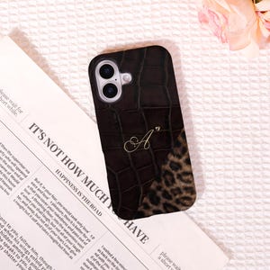 May include: A brown phone case with a crocodile skin effect, a leopard print section, and a gold letter "A". The case is designed for a smartphone and has a camera cut-out.