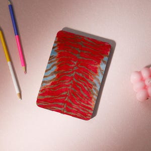 Può includere: Una copertina per quaderno con un motivo tie-dye rosso e blu. La copertina è realizzata in un materiale morbido e testurizzato.