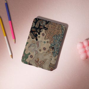 Può includere: Un taccuino rettangolare con un design patchwork nei toni del beige, blu navy e verde acqua. La copertina presenta motivi floreali e paisley. Due matite, una gialla e bianca, l'altra rosa e blu, sono posizionate vicino al taccuino. È presente anche una piccola candela rosa.