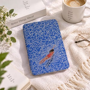 May include: A rectangular e-reader case featuring a vibrant blue and white abstract water pattern with a graphic illustration of a swimmer in a red swimsuit. The case is positioned on a textured white fabric surface near books and a coffee mug.