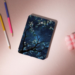 May include: A rectangular case featuring a dark blue illustration of a tree with white and blue flowers. The tree's branches extend across the case against a dark blue background. The case is on a pink surface with pencils and a candle.