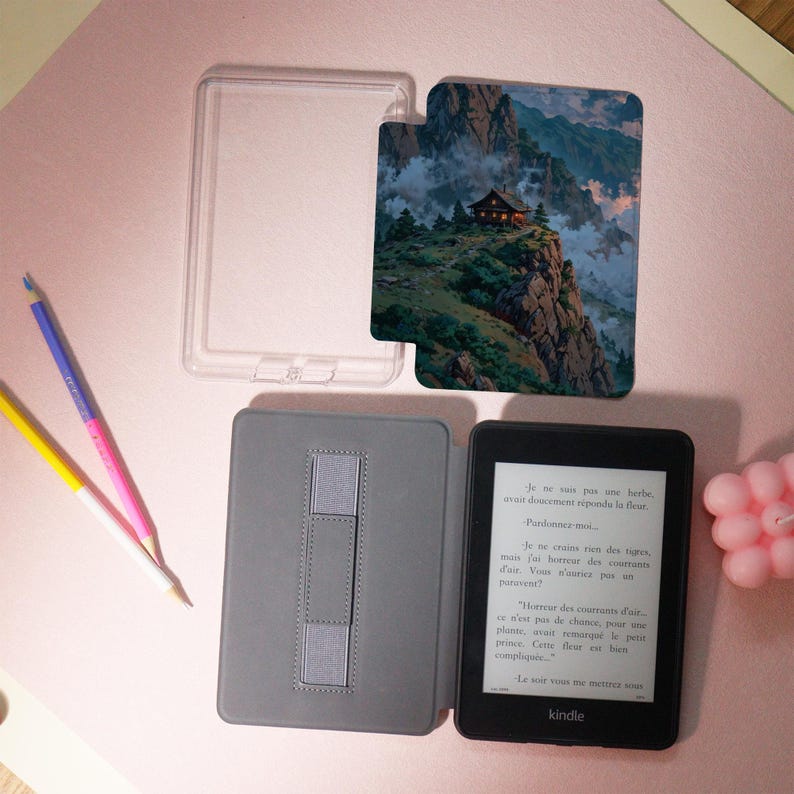 May include: A black Kindle e-reader with a gray cover and a clear plastic case. The cover features a mountain landscape design. The e-reader displays text in French. Colored pencils and a pink candle are also present.