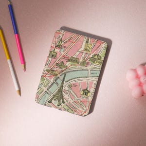 May include: A pink and cream-colored notebook featuring a vintage map of Paris, France. The Eiffel Tower is prominently displayed. The notebook is rectangular and lies on a pink surface, with colored pencils nearby.