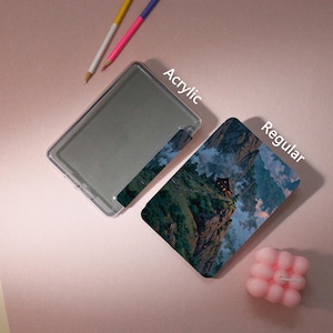 May include: Two pencils and a pink candle sit on a pink surface next to a clear acrylic case and a regular case. The acrylic case has the word "Acrylic" printed on it. The regular case has a mountain landscape design and the word "Regular" printed on it.