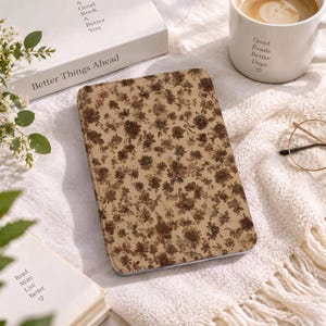 May include: A rectangular e-reader case featuring a dense, repeating pattern of dark brown flowers on a light tan background. The case is positioned on a textured white fabric surface alongside a white ceramic mug with the text Good Reads Better Days.