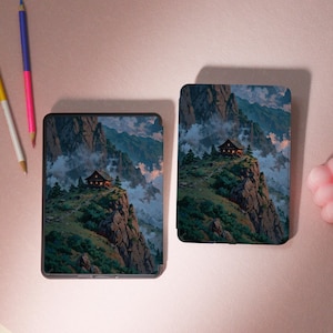 May include: Two electronic device cases with a mountain landscape design. The cases feature a small cabin perched on a cliff, surrounded by trees and mountains. The artwork is in shades of green, brown, and blue.