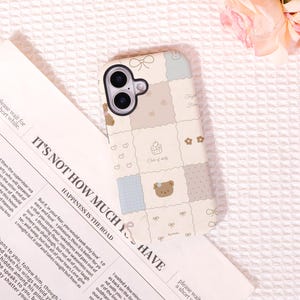 Puede incluir: Una funda para smartphone con un diseño de cuadrícula de patchwork en colores pastel suaves. El patrón incluye ilustraciones de un oso pardo, un cupcake con el texto Cake of daily, corazones pequeños, flores y lazos. La funda tiene un recorte negro para la cámara.