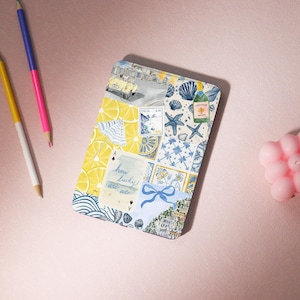 Può includere: Un taccuino rettangolare in stile acquerello con un collage di illustrazioni a tema italiano. Il design include limoni, conchiglie, un'auto d'epoca e il testo "how lucky are we". Matite e una candela rosa sullo sfondo.