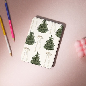May include: A rectangular, off-white notebook with a repeating pattern of green Christmas trees and beige ribbons. The notebook is on a pink surface, with three coloured pencils and a pink candle nearby.