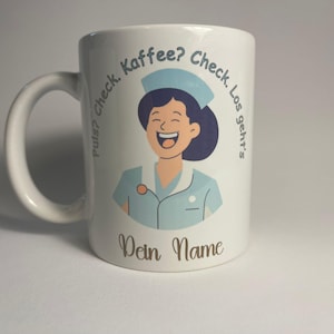 Op de afbeelding: Witte keramische mok met een cartoonillustratie van een lachende verpleegster in een blauwe uniform en een witte hoed. De tekst rond de afbeelding luidt "Pulse? Check. Kaffee? Check. Los geht's." en "Dein Name" onderaan.