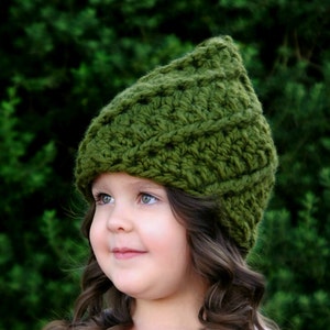 May include: A young girl wearing a green crocheted hat with a unique stitch pattern. The hat has a pointed top and covers her ears. She is wearing a pink shirt and has long brown curly hair.