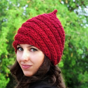May include: A woman wearing a red knitted hat with a pointed top. The hat is made with a textured stitch pattern. The woman has long brown hair and is wearing a black jacket.
