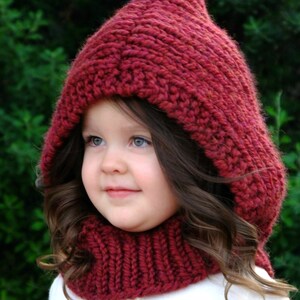 May include: A young child wearing a red knitted hooded hat and matching scarf. The hat has a pointed top and the scarf is a cowl style. The child is looking at the camera and smiling.