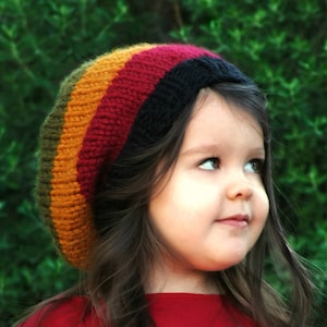 Puede incluir: Una niña pequeña con una camiseta roja y un gorro de punto de rayas de colores. El gorro tiene rayas verdes, amarillas, rojas y negras.